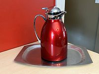 Red stainless steel carafe with stainless steel lid and spout, placed on an aluminum serving tray with rounded corners on a wooden table against a red wall.