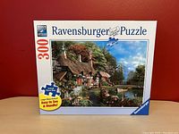 Photo of the front of the Ravensburger puzzle box showing the cottage on lake image, 300 pieces indicated, large piece format, made in Germany.