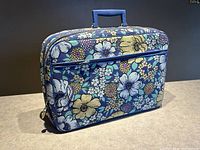 Front and side view of a rectangular retro floral suitcase with zipper and blue handle on top, showing the vibrant flower patterns and overall exterior condition.