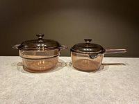 Two amber glass cookware pieces with lids on beige countertop against brown background. Shows the complete set of two cookware items with their lids.
