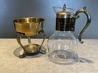 Photo showing the glass coffee carafe with silver plated handle and lid alongside the silver plated warmer stand with three legs and a space for a warming candle.