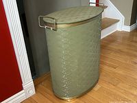 Oblong vintage hamper standing on hardwood floor near stairs showing embossed green vinyl exterior, brass side handle, and brass trim.