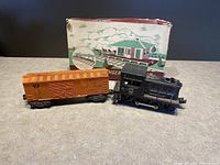 Photo of a rust-orange freight boxcar and black U.S. Army locomotive, both vintage Lionel toys, next to a vintage train station kit box.