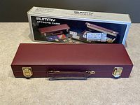 Closed maroon attaché case with gold clasps and handle, original box behind it showing product name and contents.