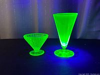 Two vintage uranium glass dessert bowls glowing neon green under UV light, one tall and one shorter with textured pattern