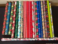23 tubes of vintage wrapping paper rolls displaying assorted holiday and seasonal patterns including florals, Santas, stripes, and characters, laid out side by side.