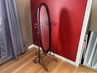 Oval cheval floor mirror with cherry wood frame, wrought iron base with curled details and bottom rack shelf.