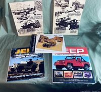 Three Jeep automotive books arranged on a green fabric background. Two large books are upright showing black and white Jeep images. The third and fourth books lay flat showing the front covers with photos of Jeep vehicles.