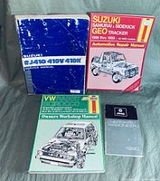 Four automotive manuals placed on a grey fabric background: blue Suzuki SJ410/410V/410X Service Manual with wear, red Suzuki Samurai & Sidekick Geo Tracker 1986 thru 1993 Repair Manual, VW Rabbit & Scirocco Owners Workshop Manual with green and white illustrated cover, and silver 1990 Jeep Cherokee/Wagoneer manual.