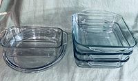 Nine clear glass baking dishes arranged on a cloth surface: four rectangular with handles stacked together, three square with handles stacked together, one square without handles, and one round dish.