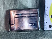 Front side of the cigarette case showing textured geometric pattern and emblem with ruler for scale.