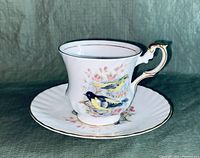 Side angle showing delicate bird illustration on white porcelain cup with ornate gold trimmed handle and matching saucer.