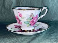 Side view of porcelain cup on matching saucer with pink rose floral design and elegant gold trim.