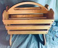 Front and top angled view of a solid wooden magazine rack showing handle, slatted design, and legs