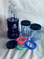 Photo showing Magic Bullet single shot blender base unit with three transparent plastic cups with black lids, one cup with blade assembly attached, and three colored plastic rings (red, blue, green) laid out in front of the black base unit.