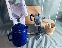 Photo shows a blue speckled enamel pot with lid, aluminum stovetop espresso maker, wooden mortar and pestle, metal garlic press, and white plastic vegetable slicer.