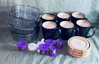 Photo showing 4 clear glass bowls stacked, 6 black mugs with textured surface, 6 purple reusable coffee pods, a white scoop, and a set of 4 round wooden coasters with holder