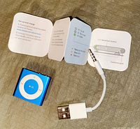Photo of blue Apple iPod Shuffle, white charging cable, and instruction manual detailing setup and battery instructions.