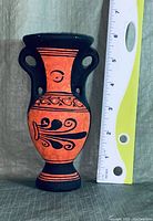 Front view of the small red and black decorative vase next to a ruler showing about 6 inches tall, with ornamental swirl patterns on the front side.