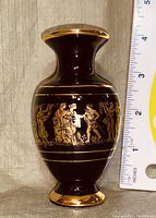Side view of a small black vase with gold classical figures and measuring ruler showing size approx 4.5 inches tall.