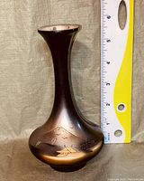 Front view of small black vase with gold engraved landscape design beside a ruler for size reference.