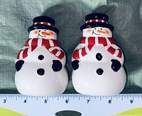 Front view of two snowman salt and pepper shakers placed on fabric with a ruler showing size about 4 inches tall.