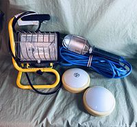 Photo showing a yellow metal-frame work floodlight, a silver handheld inspection lamp with blue cord and metal cage, and two small white dome lights with yellow bases.