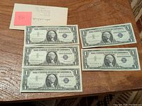 Five one dollar silver certificates from 1957 and 1957A displayed on a wooden surface with a note.