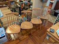 Four blonde wood mid-century style side chairs arranged in a row on a wooden floor, showing overall condition and style.