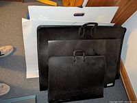 Three black artist portfolios of different sizes arranged on floor with large sheets of paper inside or nearby.