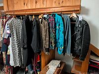 Photo showing a collection of colorful women's clothing hanging on wooden rack, including jackets and blouses with various patterns and textures.