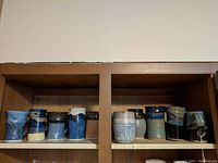 Front view of 10 ceramic mugs displayed on shelf showing different glazes, shapes, and some marked with Uchill Simons. One mug shows chip.