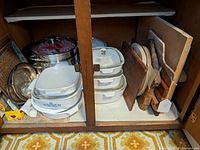 Wide view inside cupboard showing stacked Corningware Blue Cornflower pots, lids, two square serving plates, large metal double pot, and wooden cutting boards arranged vertically.