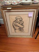 Full view of framed drawing leaning on floor with dishware behind it, showing entire artwork and frame.