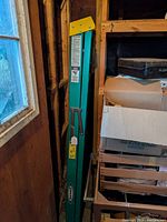 Folded green fiberglass Werner ladder leaning in corner beside window and shelf.