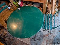 Green steel round folding bistro table shown with scratches and paint chipping, paired with matching folding chairs in a storage area.