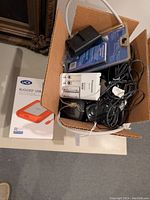 Box containing Panasonic battery charger, multiple power adapters and black cables, and the LaCie Rugged 1TB external drive box visible on side