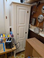 Tall white cupboard standing next to a table with a toaster and other kitchen items. The cupboard is closed, showing the paneled door and knob.