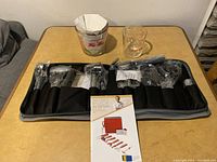 Image showing the black carrying case partially unzipped with 12 garnishing tools in plastic wrapping, alongside two measuring cups and the product leaflet on a table.