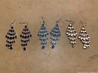 Three pairs of Avon Signature Collection dangle earrings in black, blue, and white colors laid out on a beige surface.