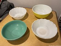 Four bowls on wooden table: two white textured milk glass, one yellow Pyrex, one teal blue bowl