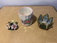 Three candle holders on a beige table: porcelain floral bouquet, ceramic cylindrical with painted flowers, and gold framed mirror holder