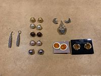 Front view of various costume jewelry including stud earrings of multiple colors, crescent moon-shaped earrings, brooches, and orange circular studs.