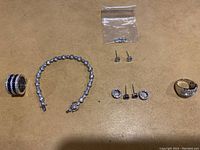 Full set laid out on tan surface showing two rings, a tennis bracelet, and six pairs of stud earrings, some with clear stones and others including blue stones.