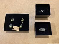 Photo of three jewelry items in black boxes: green faux pearl drop earrings with Heidi Daus tag, and two rings - one with a large clear stone and the other a wide crystal band.