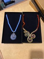 Two necklaces in presentation boxes: light blue beaded necklace with round crystal pendant on left, red-orange beaded necklace with swirl crystal pendant on right