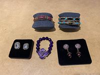 Wide view of all jewelry items including two bracelets on stands, two pairs of earrings on black pads, and purple beaded bracelet.