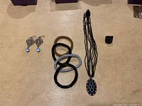 Image showing five textured bracelets in different metallic finishes, black multi-strand beaded necklace with oval pendant, and a carved black ring.