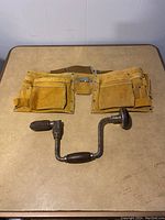 Leather nail bag and vintage brace and bit hand drill laid flat on table, showing multiple pockets on bag and overall tool condition.