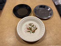 Three pie plates on a wooden surface: one white ceramic with floral design, one silver metal, one black perforated metal.
